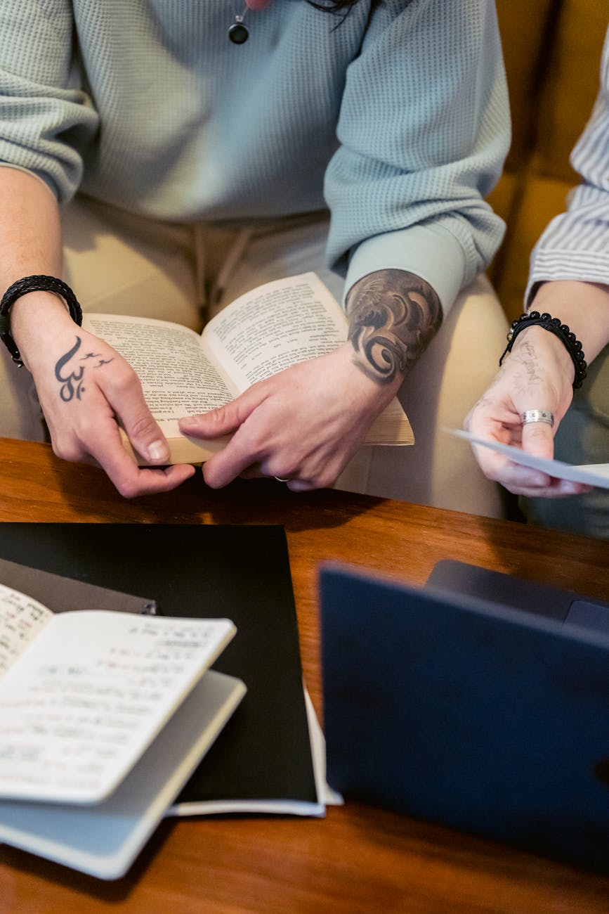 From above of crop unrecognizable tattooed person sitting on sofa and reading interesting book near friend working remotely on laptop