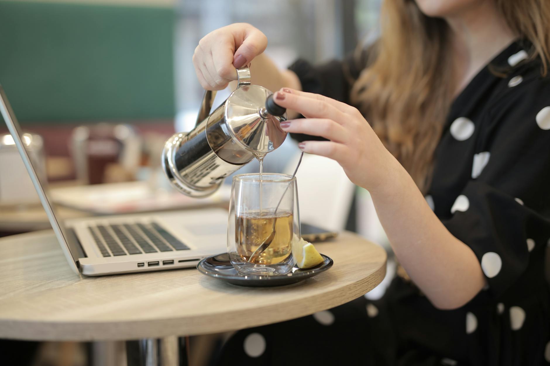 A woman pours a beverage from a French press beside a laptop in a cozy café setting.