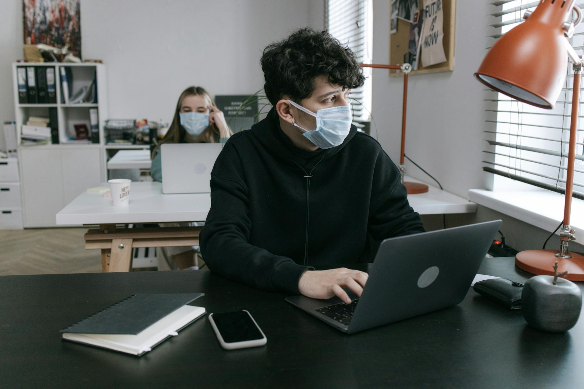 Masked colleagues working on laptops in a socially distanced office setting.