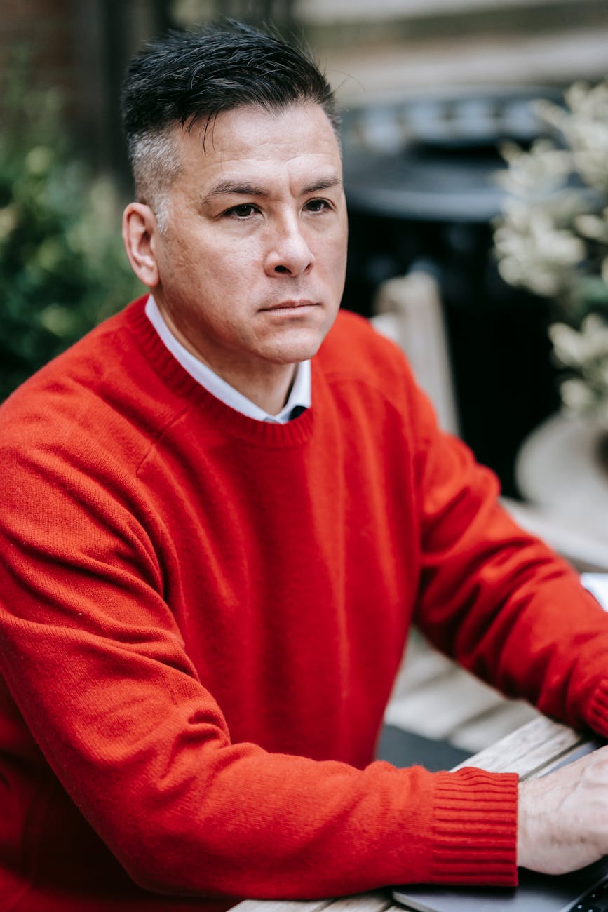 Portrait of a serious-looking adult male wearing a red sweater, sitting outdoors in a casual setting.