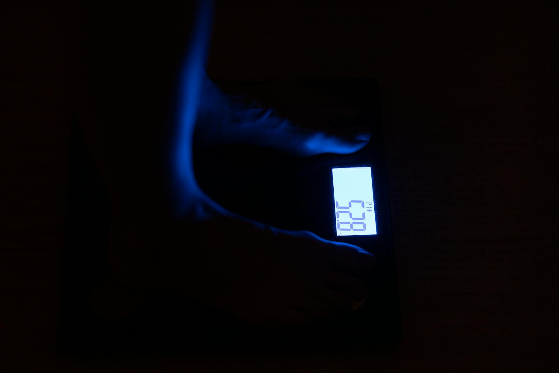 Close-up of a digital weighing scale displaying weight in a dark room with blue illumination.