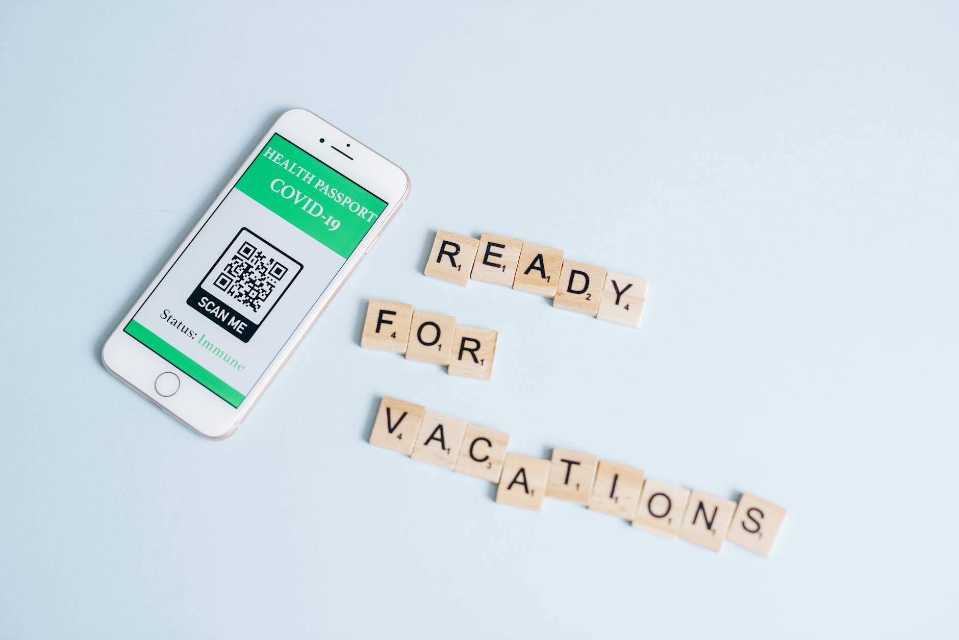 Smartphone displaying COVID-19 health passport next to Scrabble letters spelling 'Ready for Vacations' on a pastel background.