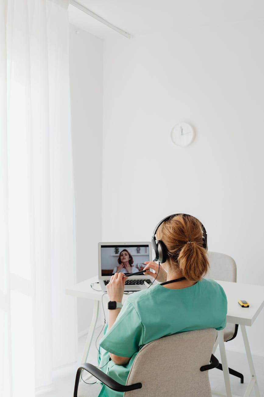 A healthcare professional in scrubs having a video call with a patient, using a laptop indoors.