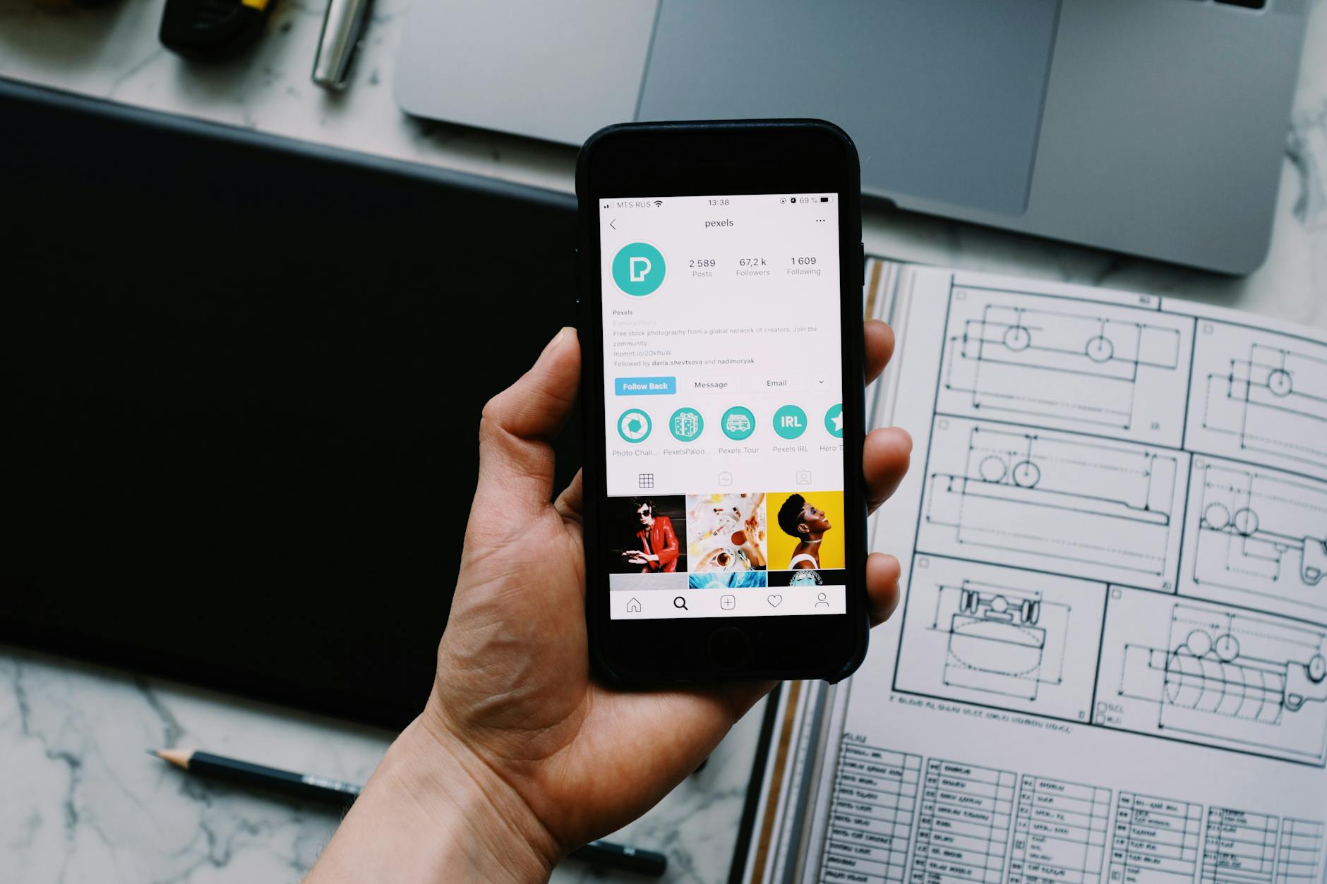 Hand holding smartphone displaying social media profile with tech gadgets on desk.