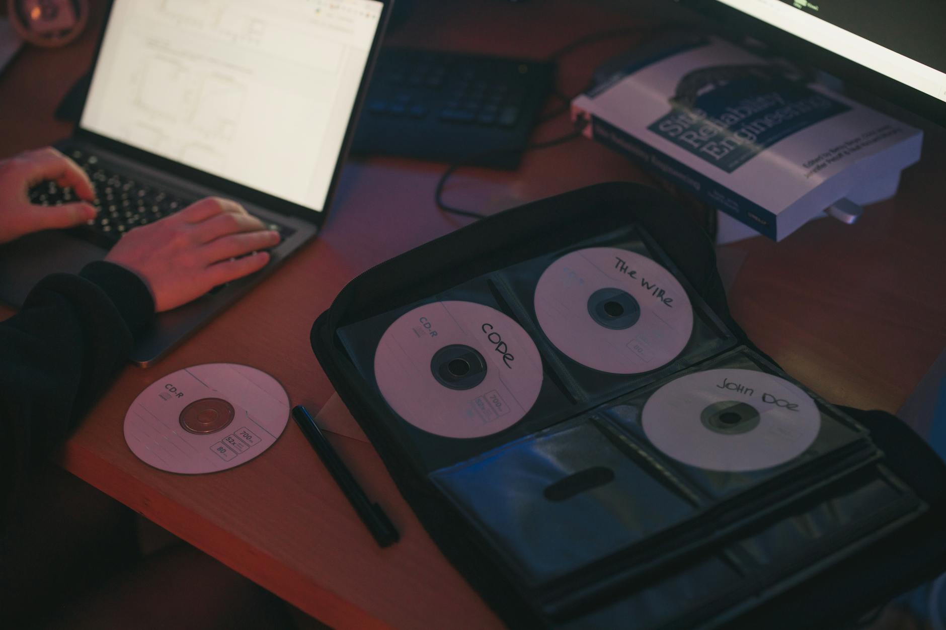 A person works on a laptop beside a set of labeled CDs on a desk.