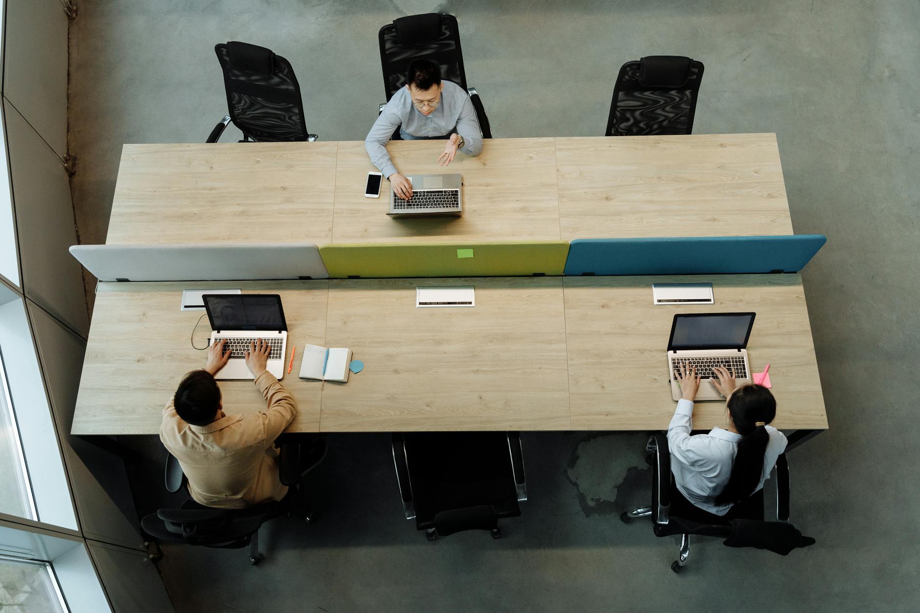 Three colleagues working at a shared office desk, using laptops in a modern workspace.