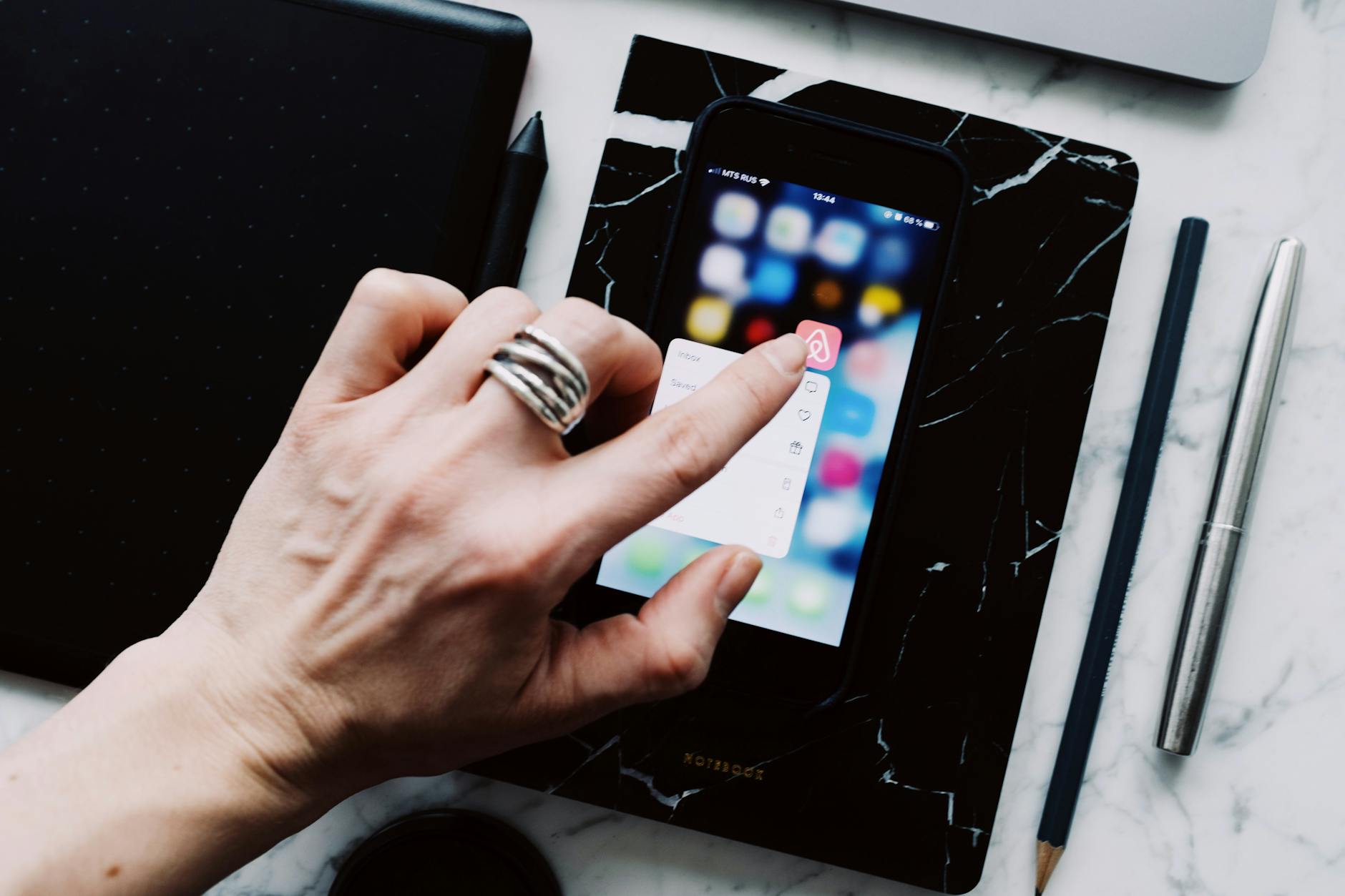 Hand interacting with smartphone on marble desk, featuring stylus and notebook.