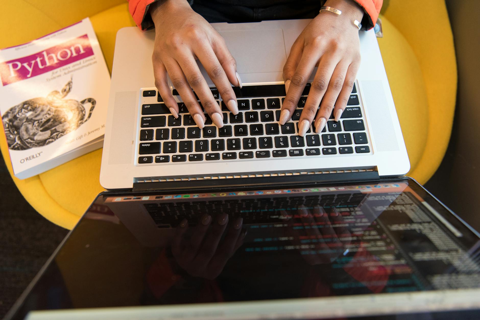Close-up of hands typing on a laptop keyboard, Python book in sight, coding in progress.