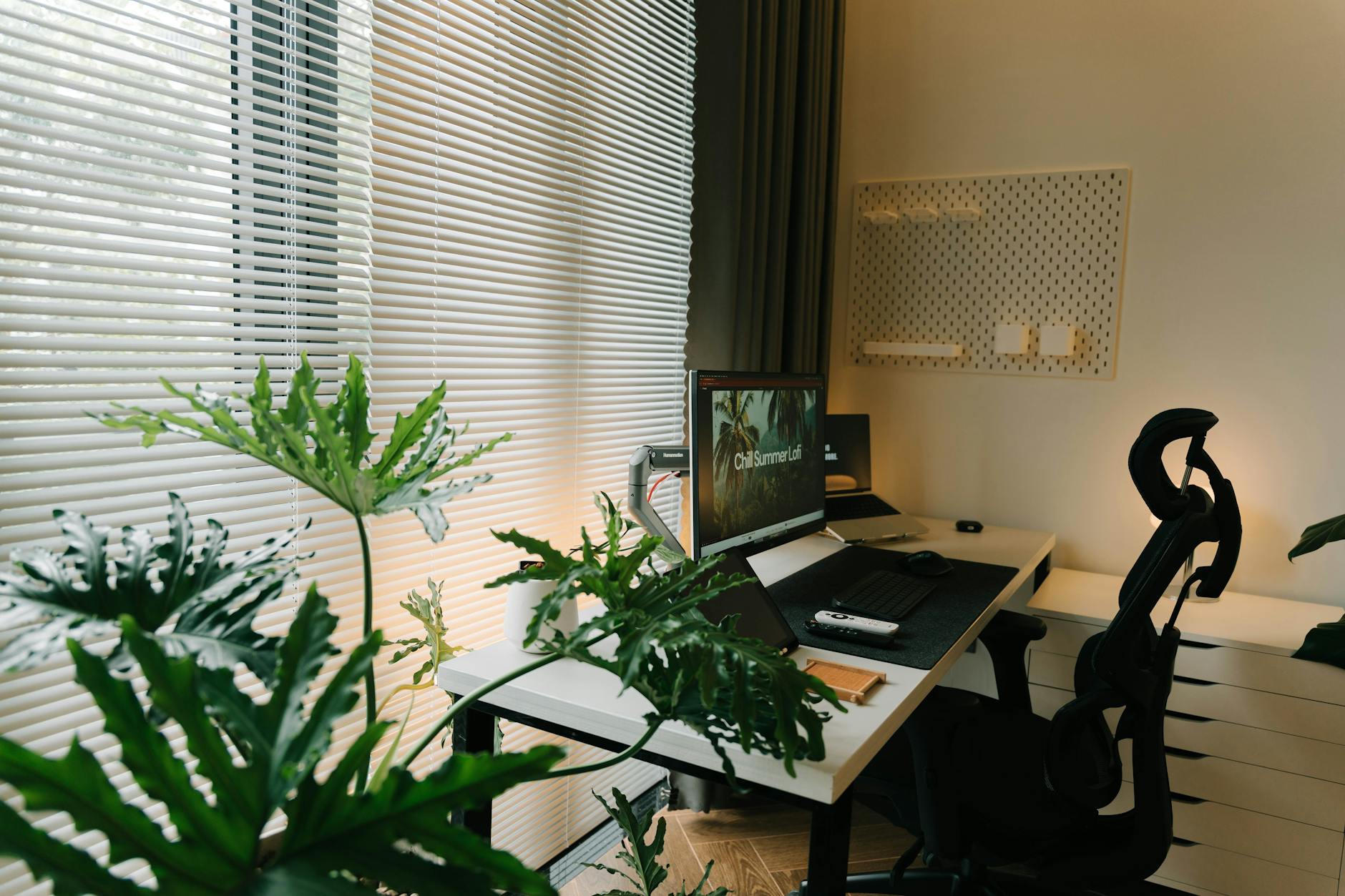 A stylish home office with a desk, chair, and lush green plants near a window with blinds.