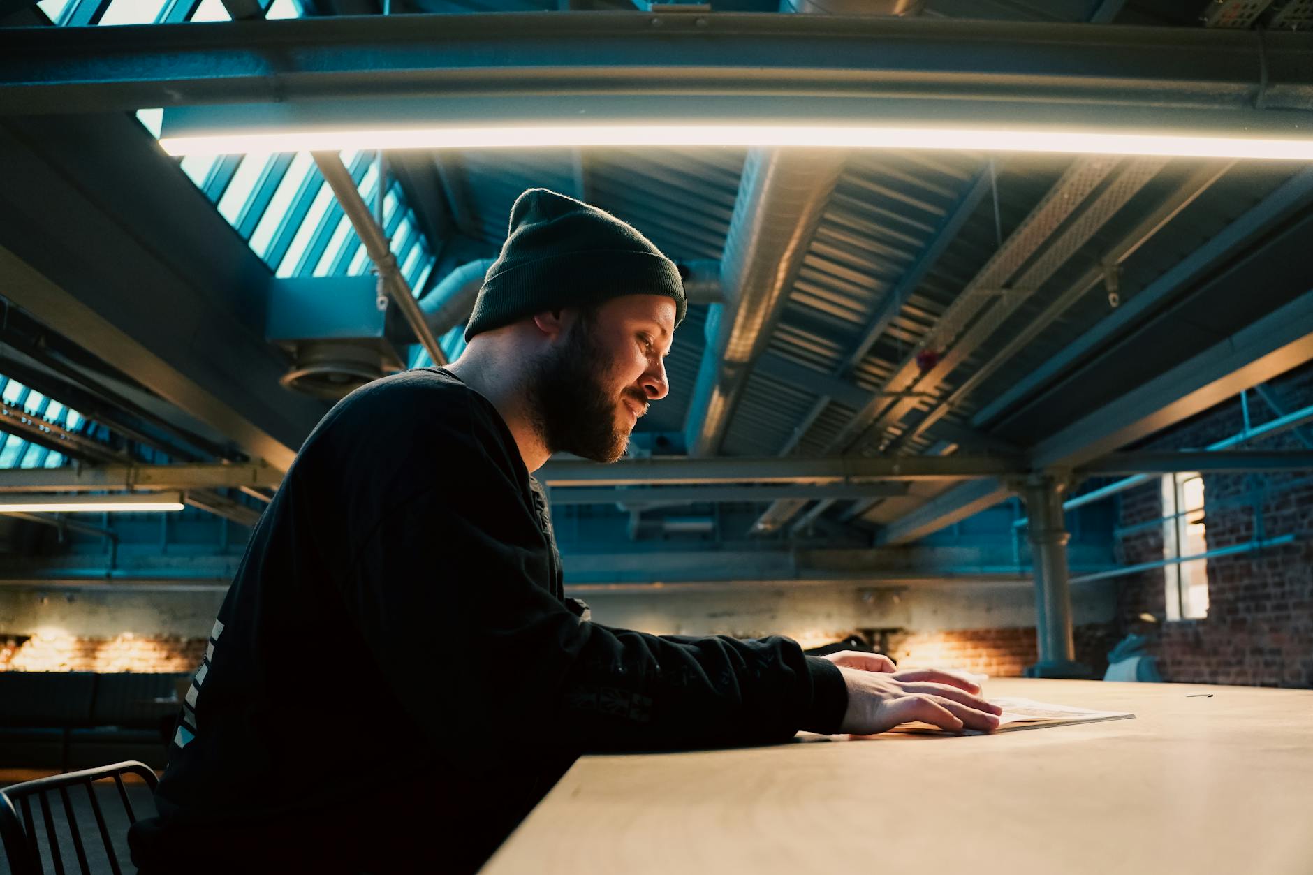 A man in a beanie reads under a modern industrial ceiling light in a loft setting.