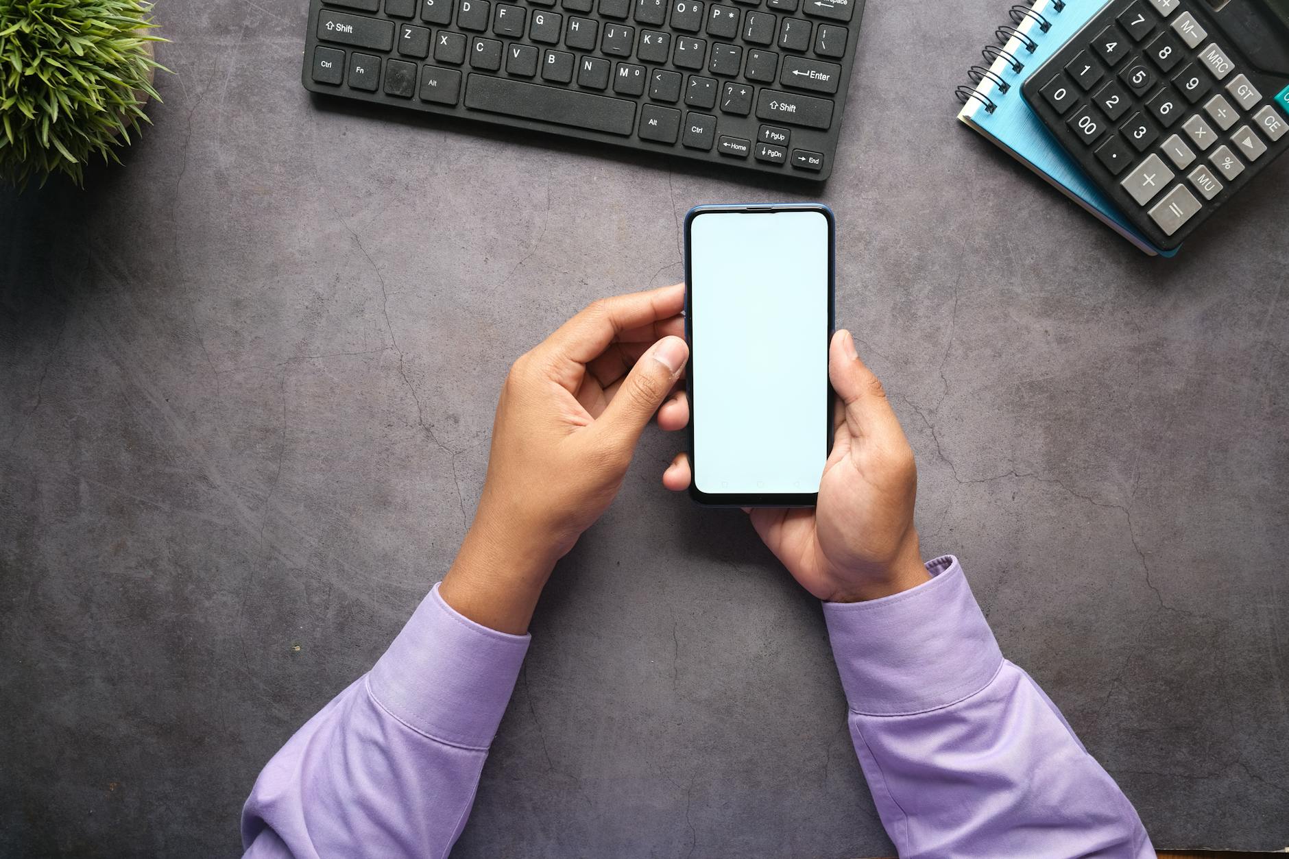 Man in purple shirt holding a smartphone with blank screen at office desk with keyboard, notebook, and calculator.