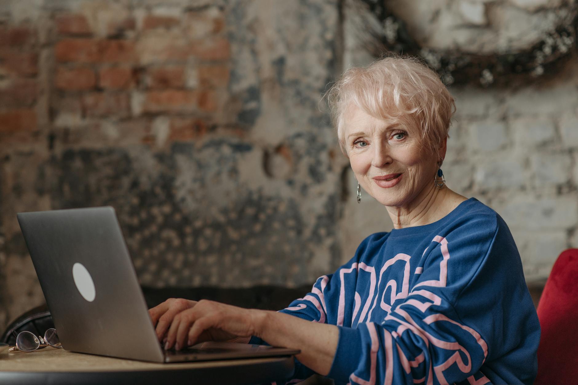 Elderly woman using a laptop at home office with rustic brick wall. A glimpse into modern senior lifestyle.