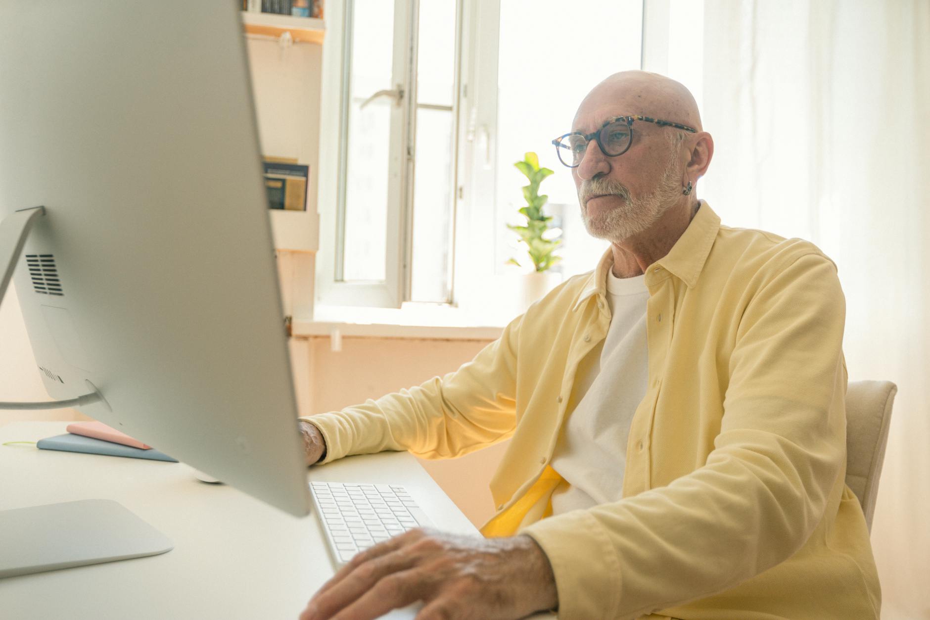 Elderly man with eyeglasses working attentively on a desktop computer indoors.