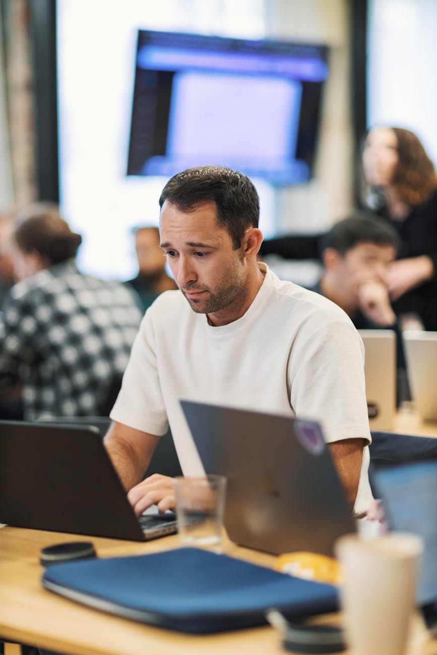 A focused adult male working on a laptop in a modern office setting, surrounded by colleagues.