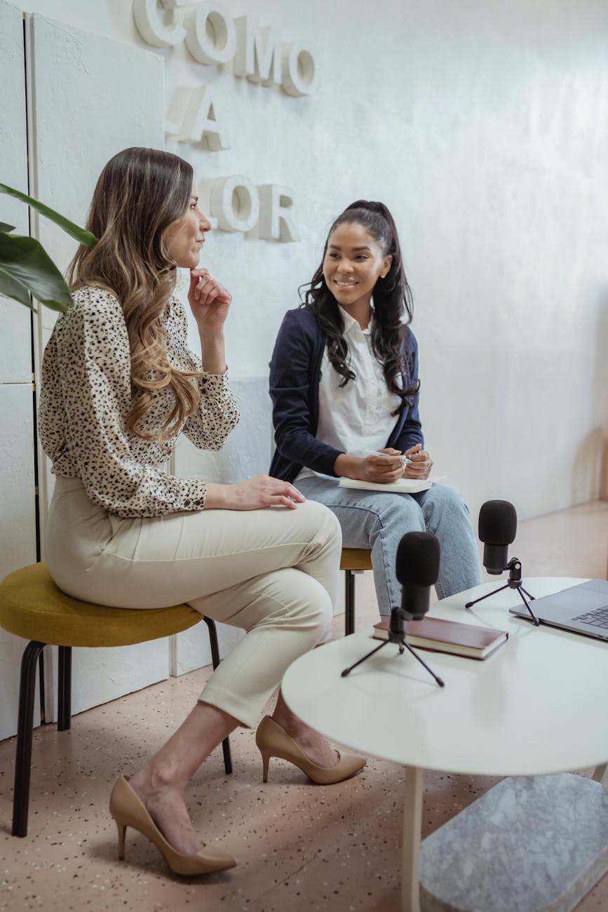 Positive multiracial female colleagues sitting on chairs near table with microphones and laptop while recording audio program in modern studio